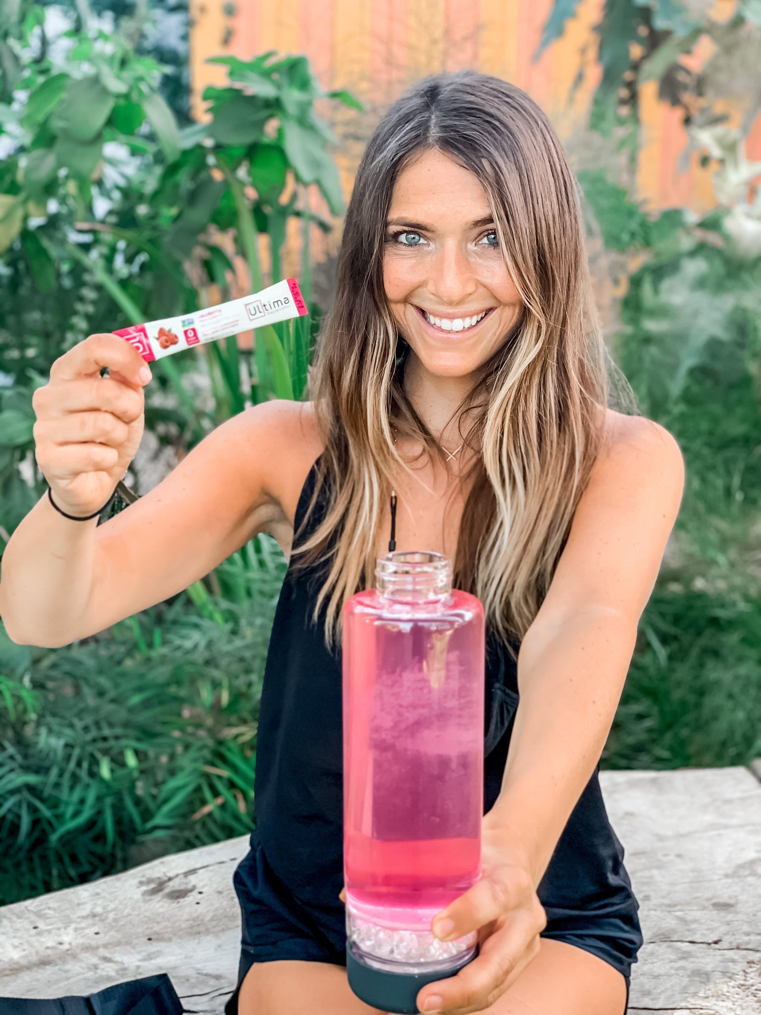 Smiling woman holds a pack of Ultima electrolytes in one hand while holding a water bottle in the other before mixing the two together for healthy hydration.