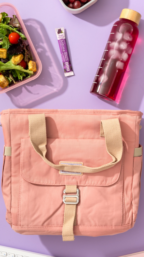 A peach-pink nylon lunch bag with beige handles and a front buckle strap, styled on a lavender background with a salad, grapes, a purple drink bottle, and a stick of protein powder.