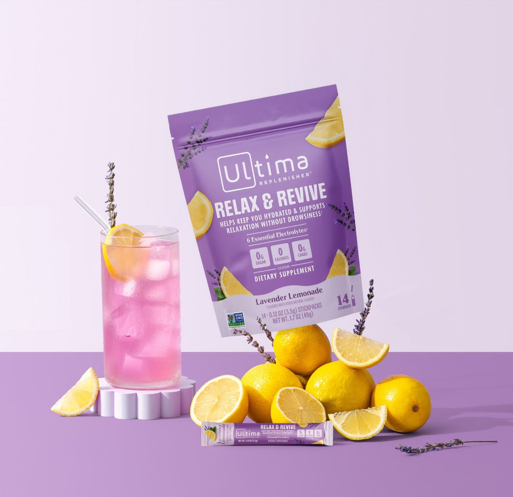 Ultima Botanicals Relax & Revive dietary supplement pouch in lavender lemonade flavor, styled with a pink drink, lemons, and lavender sprigs on a light purple background.