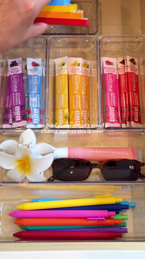 A hand organizing colorful Uline lip balm sticks into clear plastic drawer compartments. The drawers also contain a white flower clip, dark sunglasses, a pink lip balm tube, and a set of colored pens.
