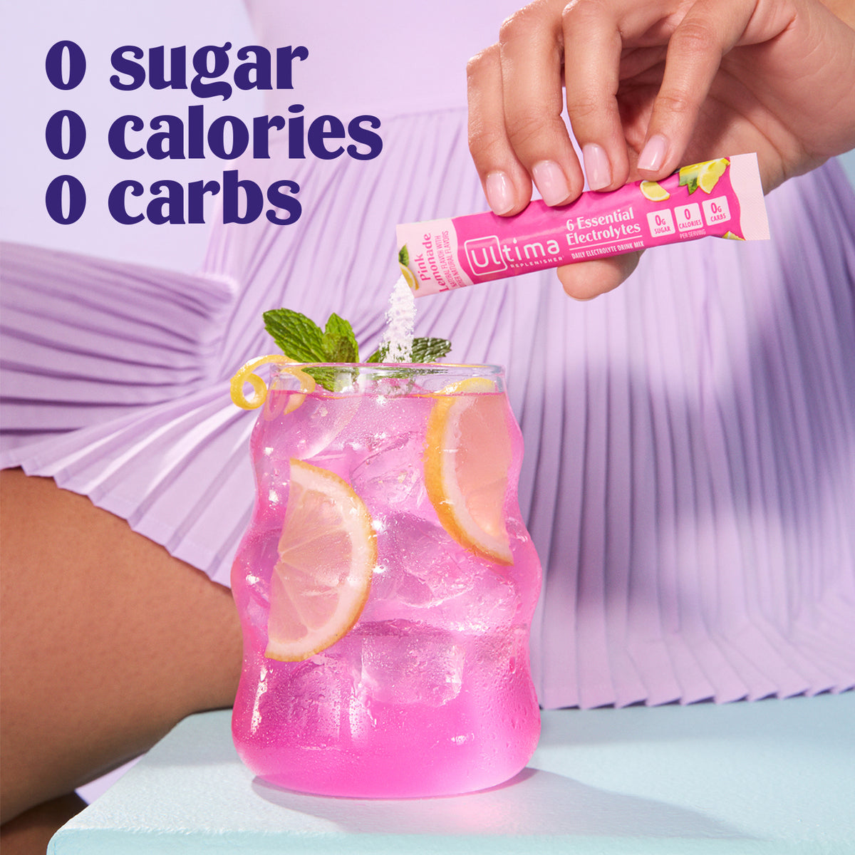 A hand pours a pink sachet of Ultimate Electrolye into a pink iced drink with lemon slices and mint, with a person in a lavender pleated dress visible in the background and text reading '0 sugar 0 calories 0 carbs'.