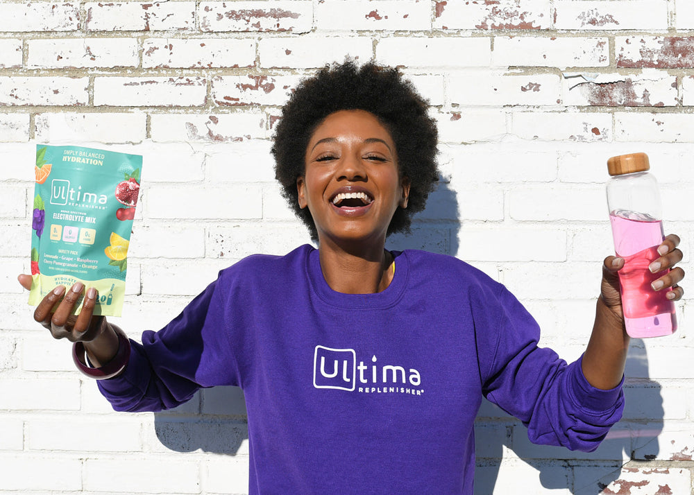 Woman holding bottle of Ultima Replenisher, stickpacks of Ultima Replenisher and wearing an Ultima Replenisher shirt